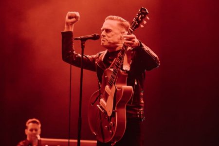 Bryan Adams cantando con guitarra eléctrica frente al micrófono en un concierto de los años ochenta.