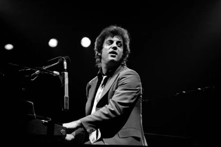 Billy Joel tocando el piano de cola y cantando con una armónica en concierto.