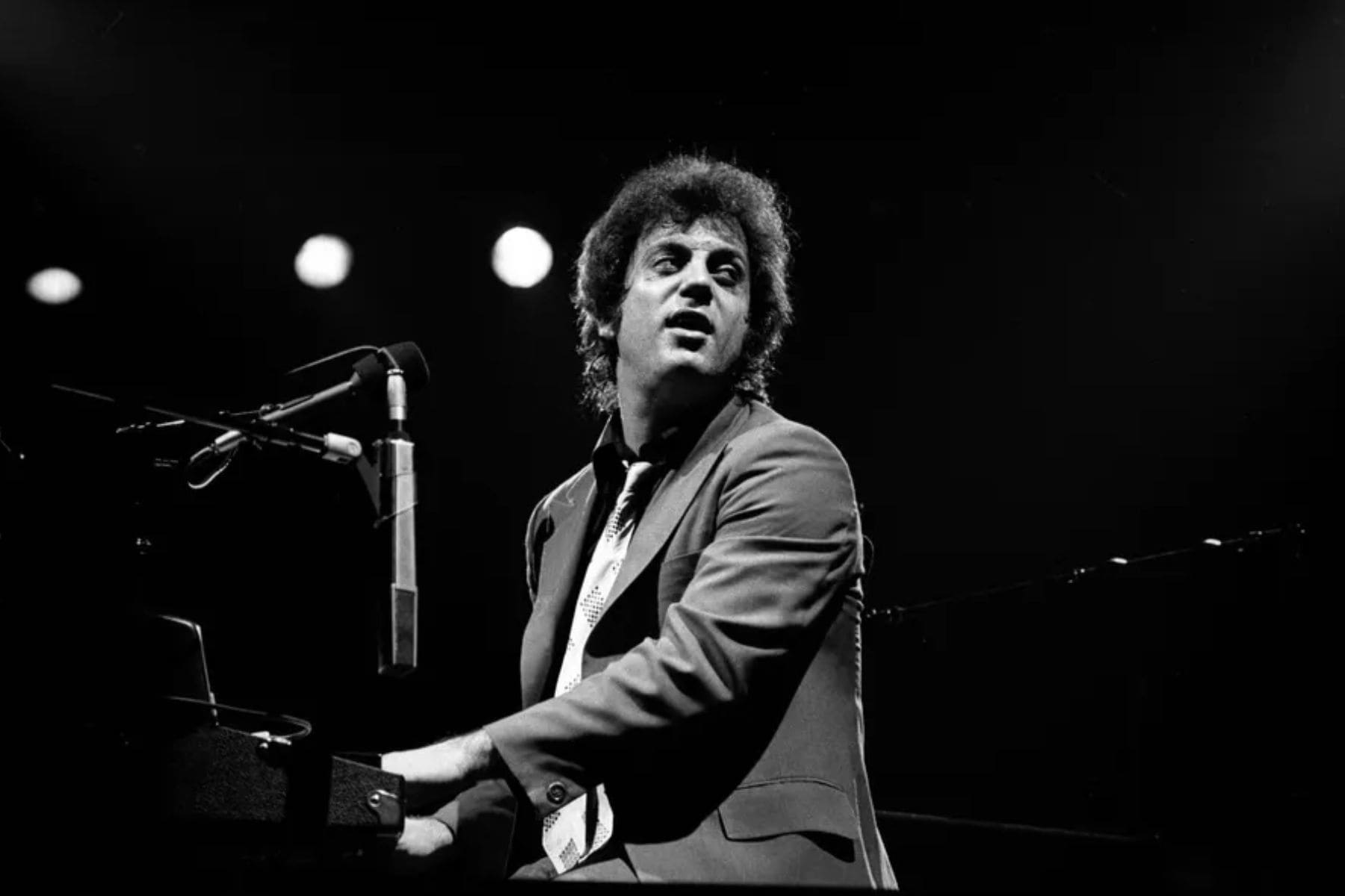 Billy Joel tocando el piano de cola y cantando con una armónica en concierto.